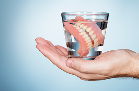 Hand holding a glass of water with dentures soaking in it